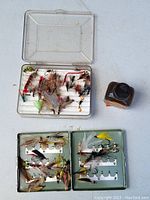 Two plastic cases containing assorted vintage fishing flies of various colors and styles, and a small bottle of oil for fly maintenance.