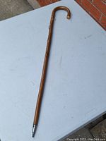 Full view of the wooden walking stick cane on a table, showing its straight shaft and curved handle, ending with a metal tip.