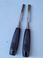 Photo showing two Sandvik chisels with black handles and flat metal blades, surface wear visible