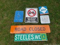 Overview of seven mixed metal signs including parking, road closed, ATV restriction, street name, and informational signs laid out on grass.