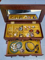 Two photos of a wooden jewelry box with a mirror containing a collection of various vintage costume jewelry pieces arranged in the compartments and drawers.