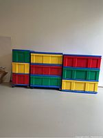 Front view of three plastic drawer bins showing multicolor drawers in green, yellow, and red with blue frame. One unit is smaller and missing wheels.