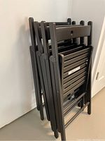 Four black wood slatted folding chairs folded and stacked against a wall.