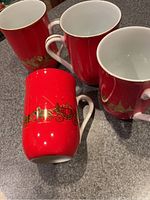 Four red mugs with white interiors and gold Christmas sleigh and tree designs, arranged on a countertop