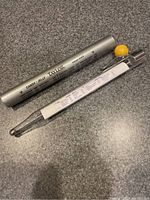 Glass candy thermometer with temperature scales in Fahrenheit and Celsius and a yellow knob, along with silver protective metal case labeled Taylor and usage instructions.