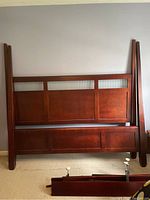 Disassembled four poster king size bedframe parts leaning against wall including headboard, footboard, rails, and posts in brown wood with ribbed glass panels.