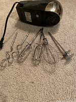 Black Oster electric hand mixer with four metal attachments laid out on carpet. Attachments include dough hooks and beaters.