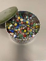 Open metal tin showing numerous vintage glass marbles in assorted colors and patterns.
