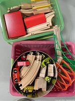 Photo showing green and pink bins with wooden railroad track pieces, several wooden toy trains in multiple colors, and plastic ramps in orange and green.