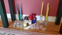 Wide view of assorted tall taper candles in blue, green, yellow, white colors, red pillar and small candles, with multiple clear glass candle holders on hardwood floor with pink wall background.