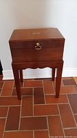 Front view of wood chest with brass handle on stand on tiled floor against white wall.