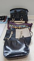Photo showing six handbags stacked; varied sizes and styles including black with crocodile texture, purple fabric with multicolor dots, and color block designs.