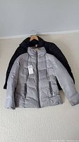 Two ladies winter jackets, grey and black, displayed on hangers on a beige carpet.