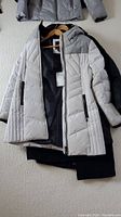Photo showing a white quilted puffer coat with zipper pocket details and lining inside, laid over a black coat on hangers.