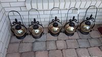 Five black metal votive patio lantern lights lined up on a stone patio against a white brick wall. Each lantern features an amber glass globe and a curved handle.