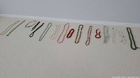 Sixteen assorted costume jewelry necklaces laid out in a row on a textured surface, showing variety in length, bead type, and color including black, green, white, red, and silver.