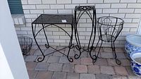 Full view of three metal plant stands with different sizes and designs placed side by side against a white brick wall