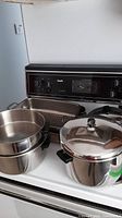 Photo showing four stainless steel pots, two rectangular roasting pans including one Paderno brand, one grill lifter placed on a white stove.