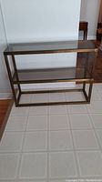 Full frontal view of the metal side table showing two glass shelves within a bronze-toned metal frame.