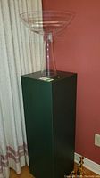 Tall clear acrylic stand with a wide bowl-shaped top, photographed against a curtain and red wall