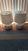 Two white ceramic table lamps with floral designs on the bases and off-white pleated drum shades placed side by side on a wooden surface in front of blinds.