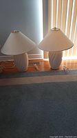 Two ceramic base table lamps with white fabric shades sitting on the floor in front of window blinds.