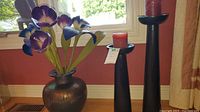Photo of metal vase with six purple and white faux flower stems beside two black tall candle holders with red candles in front of a window.