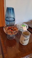 Tall blue glass vase, clear glass footed dish with potpourri, and ceramic vase with floral relief seen on a wooden surface.