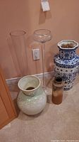 Five large vases including two glass and three ceramic placed on carpet against peach wall.