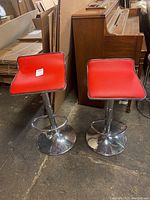 Pair of chrome bar stools with red faux leather upholstery and round pedestal bases seen from front next to wooden piano and cardboard sheets.
