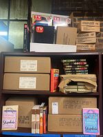 Photo showing stacked boxes of Maxell VHS tapes, TV Guide collector books, two labeled cardboard boxes of Sophie Kinsella books, and colorful hardbound children's books.