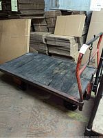 Full view of the metal framed cart with wooden planks on the platform and orange frame, situated in a warehouse environment with stacks of cardboard boxes in the background.