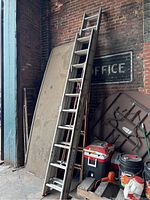 Two aluminum extension ladders leaning against a brick wall, showing height and rope pulley mechanism with nearby storage items.