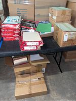 Photo showing multiple unopened and opened boxes of file folders and classification folders on a table and floor, including loose folders.