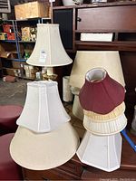 Photo showing two table lamps and six fabric lampshades stacked and placed on furniture in a warehouse setting.