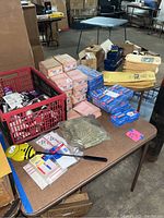 Boxes and crates of office supplies including boxed Pink Pearl erasers, note pads, coin holders, yellow store paper bags, and other stationery on a brown table in a warehouse environment.