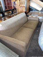 Brown upholstered sofa with three seat cushions, straight arms, viewed from front-left angle showing the side profile and seat cushions.