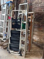 Photo showing four stepladders: one white metal painter's stepladder with three attached black plastic trays, an aluminum extension ladder in the back, and two wooden stepladders on the right side against a brick wall.
