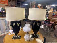 Two black wood table lamps with cream fabric shades flanking white glass electric hurricane lamp on a wooden table in warehouse setting