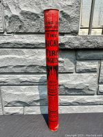 Front view of tall red tin lithographed Bickle fire extinguisher showing black printed text and graphics against a stone wall background.