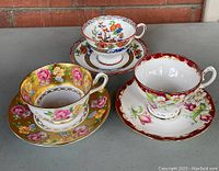 Three antique floral porcelain tea cups with matching saucers, set displayed on a gray surface in front of a brick wall.