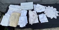 Wide view showing all the antique estate linens laid out on a black cloth, including various crocheted doilies, embroidered napkins, woven cloths, and folded white linens.
