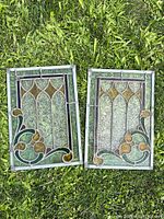 Two matching antique stained glass window panels laid on grass surface, showing full front view of each panel with colorful and textured glass design.