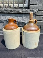 Two antique stoneware whiskey jugs, viewed front side with visible body and neck.