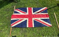 Full view of the antique cotton Union Jack flag laid flat on grass, attached to a wooden pole along the vertical side. Shows the full flag pattern and size.