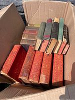 Box of antique books showing a variety of bindings, primarily red leather with gold embossing on larger volumes, and assorted smaller books with different cloth and leather covers.