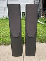 Front of pair of Mission M74i speakers showing angled black fabric grills and wood veneer sides outdoors on concrete with grass background.