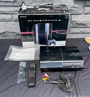Full lot setup showing Sony PS3 40GB console with remote, AV cable, manuals, and original box on black fabric.