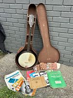 Full view of vintage Harmony banjo inside its brown hard case along with multiple vintage music books and string packages arranged outside the case.