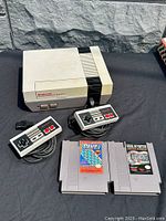 Overall view of Nintendo NES console, 2 wired controllers, Jeopardy! and Wheel of Fortune game cartridges on black cloth surface